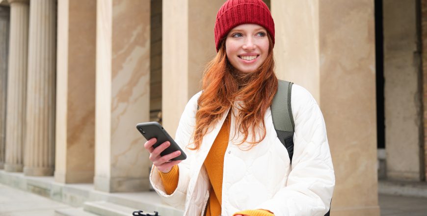 Redhead girl, tourist with backpack, uses mobile phone to rent e-scooter on streets of European city